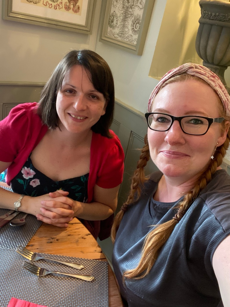 A selfie of me and my friend in a restaurant. I'm wearing a grey t-shirt with a pink headband and my friend is wearing a red cardigan over a floral top. Both of us are smiling